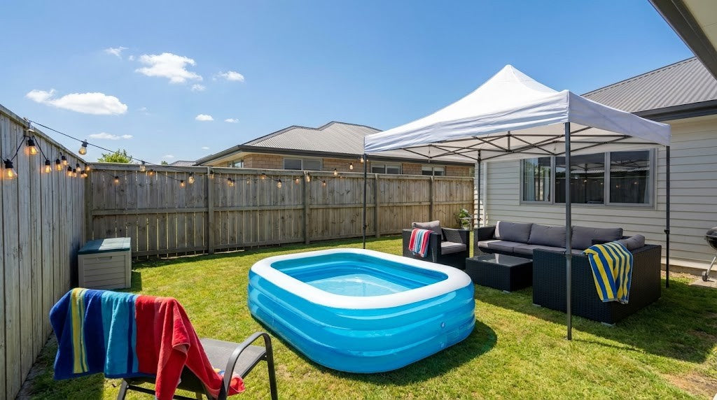 Inflatable family pool on a green lawn in a New Zealand backyard with a pop-up gazebo for shade, rattan outdoor sofa set with cushions, and festoon lights strung overhead