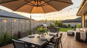 Outdoor dining table set for six under a cantilever umbrella with festoon lights overhead and a rattan lounge area in a New Zealand backyard at golden hour